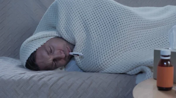 咳止めシロップと紙ナプキンの近くの毛布にソファに横たわっている間 病気の男性は温度を測定します — ストック写真