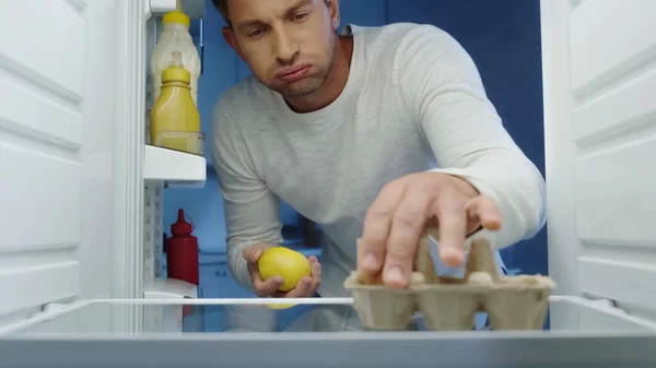 Unhappy Man Puffing Cheeks While Taking Egg Lemon Fridge — Zdjęcie stockowe