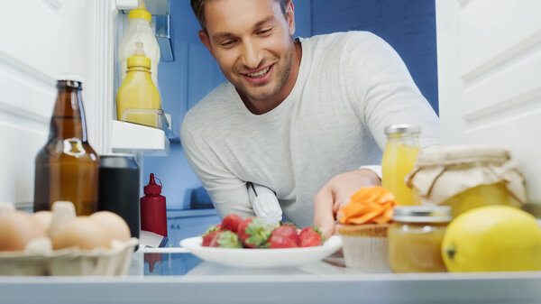 joyful man taking ripe strawberries from fridge with beverages, sauces, eggs and jars with food