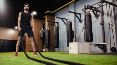 full length of bearded sportsman holding battle ropes in gym