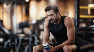 tired sportsman in earphone listening music and holding sports bottle in gym 