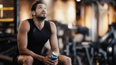 tired sportsman in earphone listening music, resting and holding sports bottle in gym 