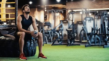 exhausted man in earphone listening music, sitting on car tire and holding sports bottle in gym