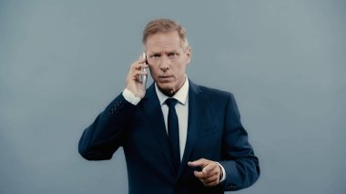 Mature businessman pointing with finger and talking on smartphone isolated on grey 