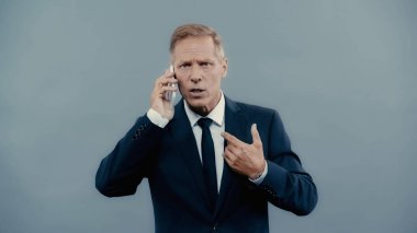 Mature businessman gesturing and talking on smartphone isolated on grey 