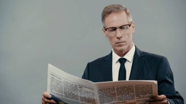 Mature businessman in suit and eyeglasses reading newspaper isolated on grey 