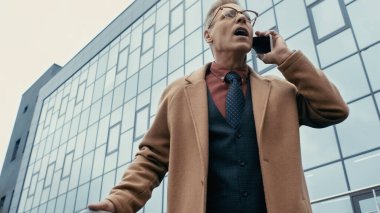 Low angle view of mature businessman in coat talking on cellphone outdoors 