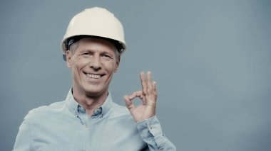 Smiling engineer in hardhat showing okay gesture isolated on grey 
