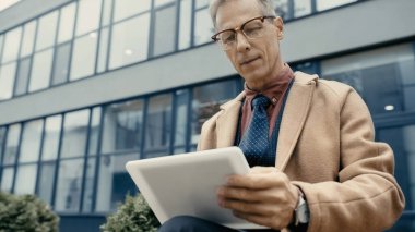 Mature businessman in coat using blurred digital tablet on urban street 