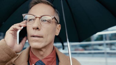 Mature businessman in coat and eyeglasses talking on cellphone and holding umbrella outdoors 