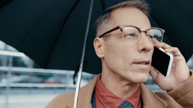 Businessman in eyeglasses and coat holding umbrella and talking on mobile phone on urban street 