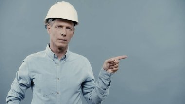 Mature engineer in helmet pointing with finger and looking at camera isolated on grey 