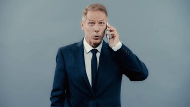 Excited businessman talking on smartphone and looking at camera isolated on grey 