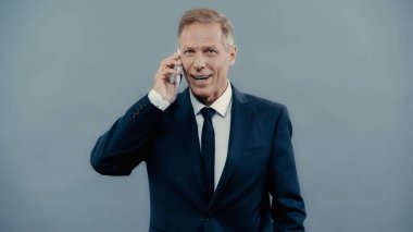 Positive businessman in formal wear talking on smartphone isolated on grey 