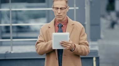Mature businessman in coat using digital tablet on urban street 