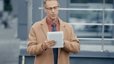 Middle aged businessman in coat using digital tablet on urban street 