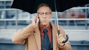 Angry businessman talking on smartphone and holding umbrella outdoors 