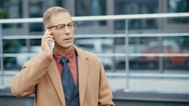 Mature businessman in coat talking on mobile phone on urban street 