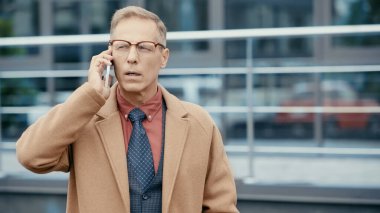 Middle aged businessman in formal wear and eyeglasses talking on smartphone outdoors 