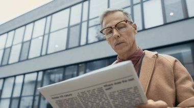 Low angle view of mature businessman in coat and eyeglasses reading news on urban street 