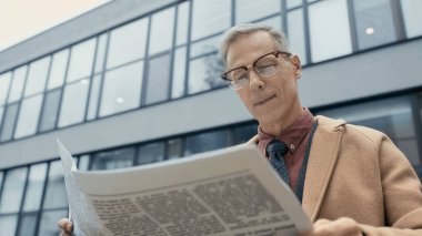 Low angle view of smiling businessman in coat reading newspaper outdoors 