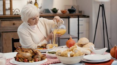 senior man pouring orange juice near roasted turkey, grilled corn and buns on table