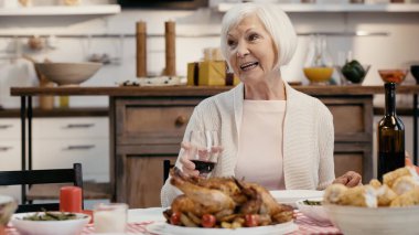 happy senior woman holding glass of red wine near traditional roasted turkey during thanksgiving dinner