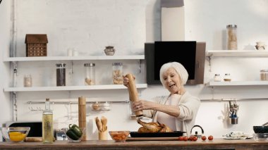 joyful senior woman seasoning roasted turkey near fresh vegetables and oil in kitchen