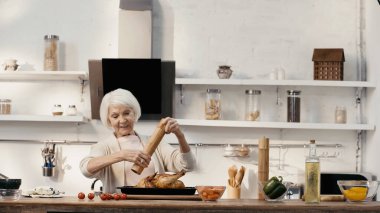 smiling senior woman seasoning roasted turkey near fresh vegetables and oil on table