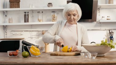 senior woman cutting bell pepper near bowls with fresh vegetables in kitchen
