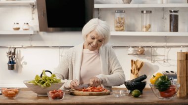 happy senior woman cutting bell pepper near fresh cherry tomatoes, lettuce and carrots