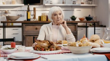 lonely and thoughtful senior woman sitting near thanksgiving dinner served on table in kitchen
