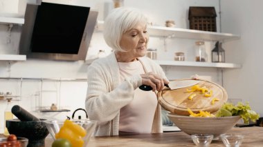 positive senior woman preparing vegetable salad and adding sliced bell pepper into bowl