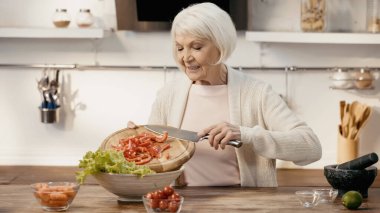 smiling senior woman adding sliced bell pepper to lettuce while preparing vegetable salad