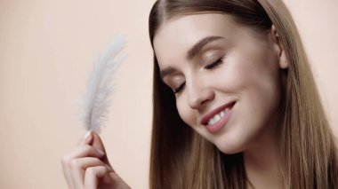 happy young woman with closed eyes holding feather isolated on beige 