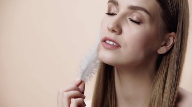 young woman with closed eyes holding feather near face isolated on beige 