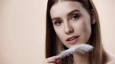 young woman holding feather near face and looking at camera isolated on beige 