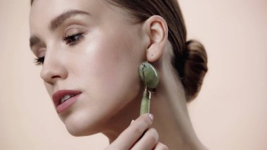 young woman looking away while using jade roller isolated on beige 