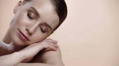 feather near cheek of pretty woman with closed eyes isolated on beige 