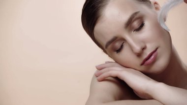 soft feather near cheek of pretty young woman with closed eyes isolated on beige 