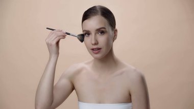 young woman with bare shoulders applying face powder while holding cosmetic brush isolated on beige
