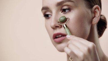 pretty young woman using jade roller while massaging and lifting face isolated on beige 