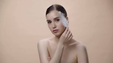 young woman with bare shoulders holding white feather near face isolated on beige 