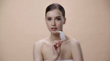 sensual young woman with bare shoulders holding white feather near face isolated on beige 