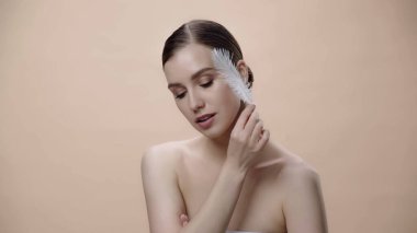 sensual woman with bare shoulders holding white feather near face isolated on beige 