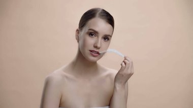 young woman with bare shoulders holding white feather isolated on beige 