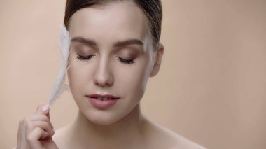 young woman with closed eyes and perfect skin holding feather isolated on beige 
