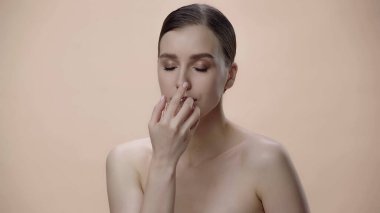 young woman with bare shoulders holding bottle while smelling perfume isolated on beige