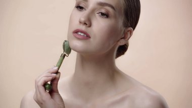 young woman with bare shoulders massaging face with jade roller isolated on beige 