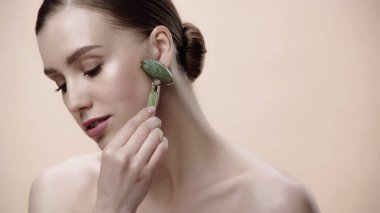 pretty woman with bare shoulders massaging face with jade roller isolated on beige 